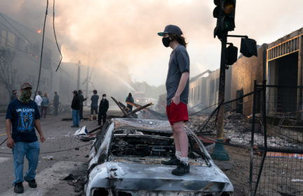 A man stands on a burned out car on Thursday morning as fires burn behind him in the Lake St area of Minneapolis, Minnesota. Photo: Lorie Shaull from St Paul, United States